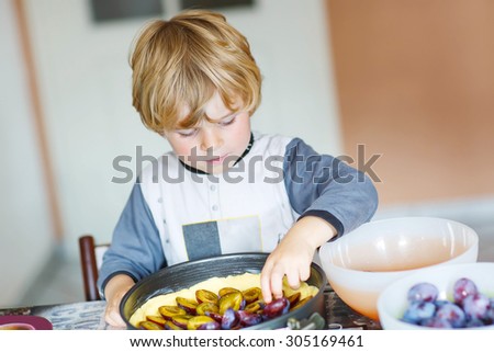 Funny cute little kid boy helping and baking plum pie in domestic kitchen, indoor. Child covering cake with fruits.