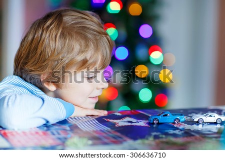 Little blond child playing with cars and toys at home, indoor. funny boy having fun with gifts. Colorful christmas lights on background. Family, holiday, kids lifestyle concept.