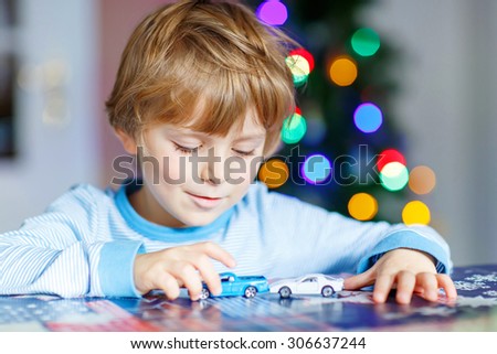 Cute blond child playing with cars and toys at home, indoor. funny boy having fun with gifts. Colorful christmas lights on background. Family, holiday, kids lifestyle concept.