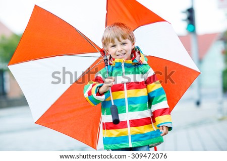 Little blond kid boy walking with big umbrella outdoors on rainy day. Child having fun and wearing colorful waterproof clothes and rain boots. Family, happy childhood,  kids concept
