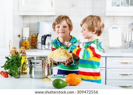 Two funny kid boys of 4 years cooking pasta and fresh vegetables in domestic kitchen, indoors. Sibling children in colorful shirts. Family, childhood, healthy food for kids concept