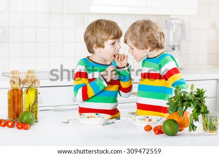 Two funny twin kids boys having fun and eating meal with spaghetti and fresh vegetables in domestic kitchen, indoors. Sibling children in colorful shirts. Family, childhood, healthy food  concept