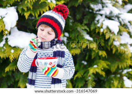 Cute little boy holding big cup with snowflakes and hot cocoa and chocolate drink and marshmallows. Kid boy in winter sweater, hat, long warm scarf and colorful gloves. On cold snowy winter day.