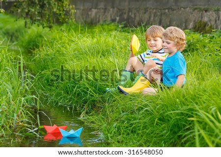 Two little brothers playing with paper boats by a river on warm and sunny summer day. Active leisure for children. Kid boys having fun together outdoors.