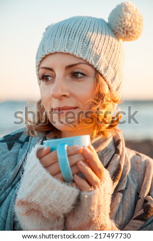 Cute young european woman in knitted clothes drinks coffee on the beach at sunset