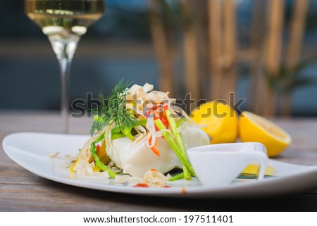 Serving of steamed sea bass fillet on a plate in a restaurant, on a wooden table with white wine and decor