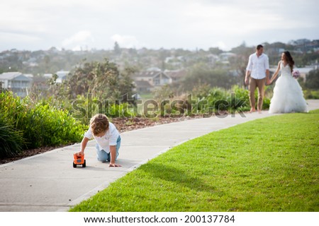 Cute toddler boy playing with his toy truck while his parents are walking along the coastline in wedding cloths on their wedding day