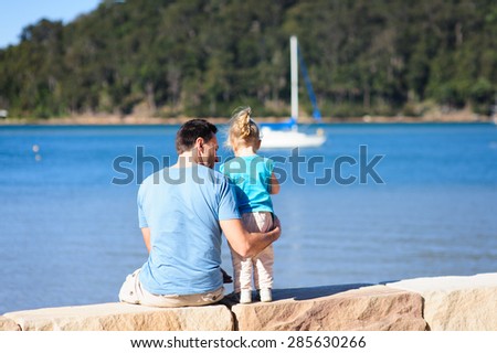 Dad hugging his child, a baby girl sitting on a rock wall with their backs to the camera on a sunny summer day with the sea and boats in the background
