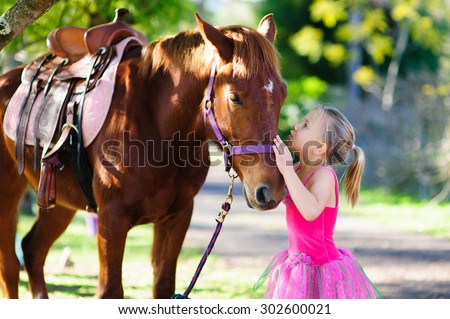 An outdoor portrait of an adorable little girl wearing pink fairy dress giving a kiss to a brown horse in a garden on a sunny summer day