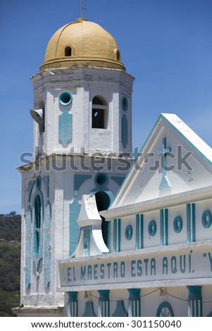 Little small blue and white small colonial church against a clear blue sky with a cross, basic decoration and letters on the facade. Merida State Venezuela
