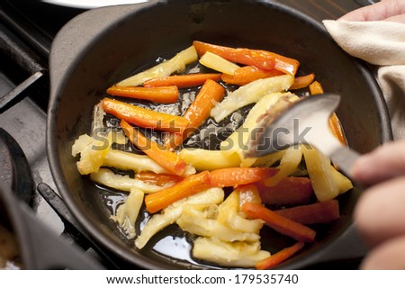 Fresh winter carrot and parsnip batons being sauteed in a metal skillet with a hand holding a spoon above them