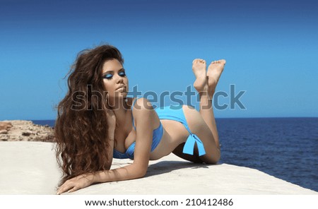 Beautiful girl model with bronzed skin sunbathing at seaside in bikini. Makeup. Long wavy hair style.