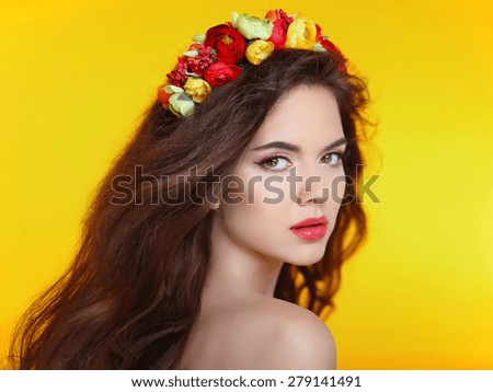 Beautiful girl model with flowers in long brown wavy hair isolated on yellow studio background. Makeup. Care. Treatment.