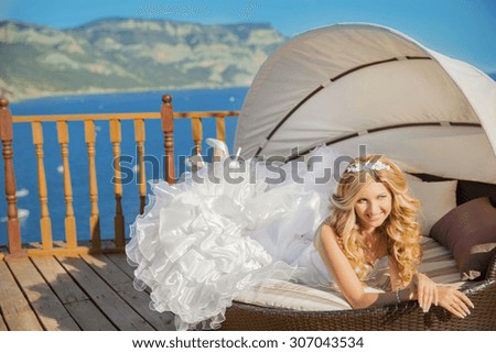 Happy funny bride in white wedding dress lying on sofa over the sea and mountains.