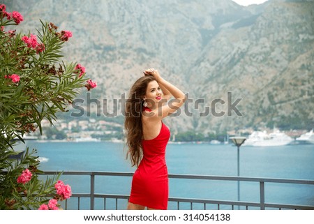 Beautiful young smiling girl enjoying over mountain and sea shore. Kotor, Montenegro. Slim Brunette model in red dress.