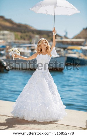 Beautiful smiling bride girl in wedding dress with white umbrella and bouquet of flowers posing on seafront, outdoors portrait