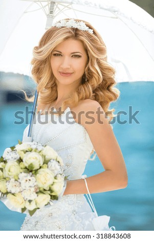 Beautiful smiling bride girl in wedding dress with white umbrella and bouquet of flowers against the seafront, outdoors portrait
