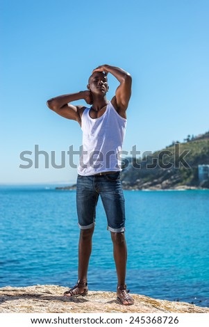 Standing African black man wearing white vest and blue short jeans. Male model thinking while isolated alone by a blue ocean and sky background. Cape Town South Africa