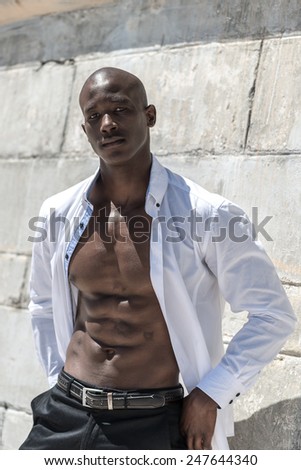 African black man model with six pack in unbuttoned white shirt, isolated against a concrete wall background on the beach