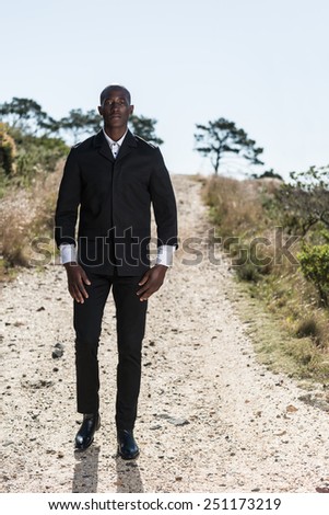 African black male model, wearing black suit with white unbuttoned shirt, standing on gravel path outdoors in nature with mountain views.