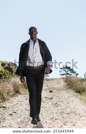 African black male model, wearing black suit with white unbuttoned shirt, walking down gravel path outdoors in nature with mountain views.