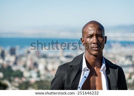 African black male model, wearing black suit with white unbuttoned shirt, standing on gravel path on a mountain view point, outdoors in nature with a city in the background