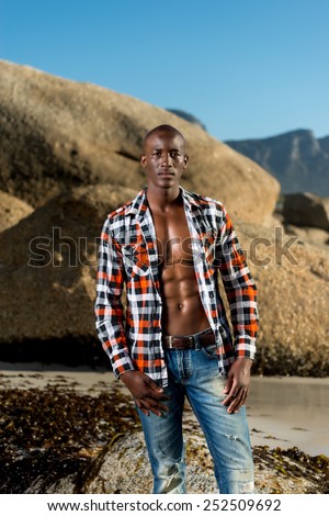 African black man model with six pack in unbuttoned checkered shirt, isolated against a beach rocks, sand and blue sky