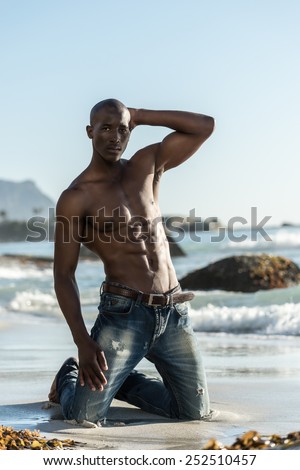 African black man model with six pack topless, isolated against a beach rocks, sand and blue sky while kneeling down during sunset