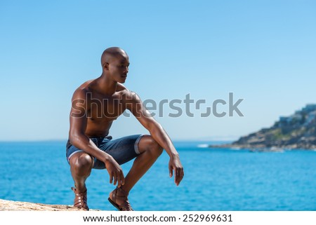 Topless African black man crouching with short jeans. Male model thinking while isolated alone by a blue ocean and sky background. Cape Town South Africa