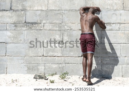 Topless, fit muscular african american male model isolated against a white concrete wall, wearing maroon shorts.