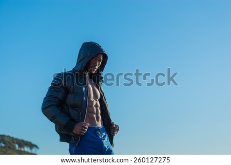 Fit, athletic and muscular African black male wearing black bomber jacket hoodie with zipper, isolated with blue sky