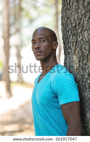 African american black man with blue shirt, standing in park, next to a tree in a park during summer