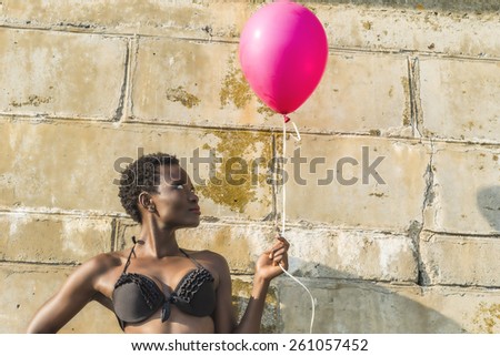 African black woman in bikini at beach, against concrete wall smiling with pink birthday balloon