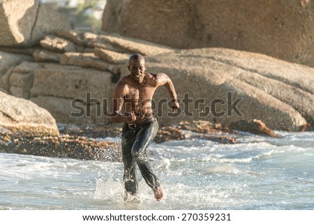 Topless, athletic, muscular and healthy black man running along the beach, splashing water during sunset