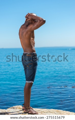 Topless African black man standing with short jeans. Male model thinking while isolated alone by a blue ocean and sky background. Cape Town South Africa