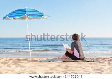 Middle-aged woman sitting on the beach reading a book. Under a beach umbrella.