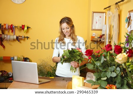 Small business owner. Portrait of mature florist woman standing at her flower shop behind the laptop while holding hands bouquet and smiling.