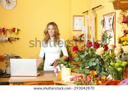 Small business owner. Portrait of mature florist standing at her flower shop behind the computer and holding hand mobile while looking at camera.
