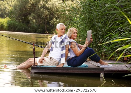 Portrait of elderly couple relaxing at nature. Senior couple sitting back to back on the pier at lake while surfing on internet. Old woman holding digital tablet in her hands.