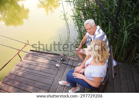 Senior couple relaxing at the lakeside. Smiling senior woman and man sitting on the pier and fishing.
