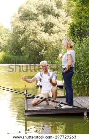 Portrait of senior couple fishing at the pier. Old man man squatting on the pier and shows to his wife how much fish caught. They are relaxing on th lakeside.
