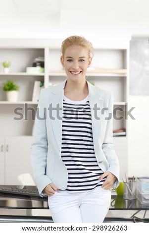 Portrait of young assistant standing at workplace in front of desk while looking at camera and smiling. Attractive businesswoman wearing casual clothing.