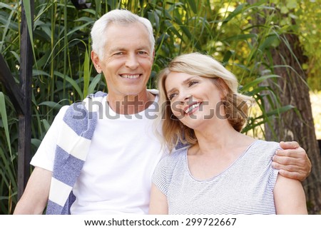 Close-up portrait of happy senior couple sitting at the lakeside. Elderly man hugs his wife while relaxing at nature.