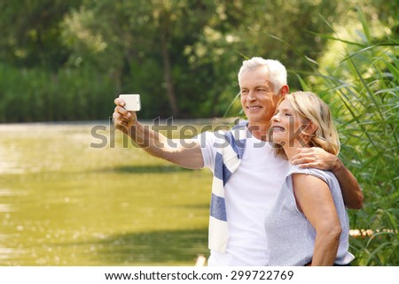 Portrait of senior couple sitting at lakeside. Elderly man hugs her wife while take self portrait with mobile.