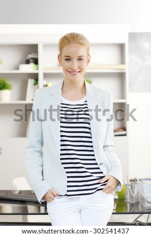 Portrait of young businesswoman standing at office in front of desk. Creative female wearing casual clothing and smiling while looking at camera.