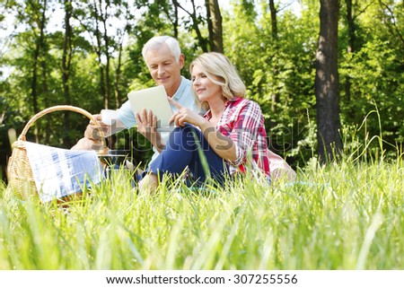 Portrait of senior hikers relaxing at forest. Elderly woman and old man sitting in the grass on blanket while using digital tablet to read online map.