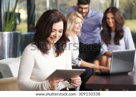 Portrait of mature businesswoman sitting at office and checking e-mails on digital tablet while her colleagues working at background in front of laptop. Teamwork at office.