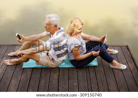 Portrait of senior couple sitting facing each other on the pier at the lake. Elderly woman holding hand digital tablet while old man using mobile phone.
