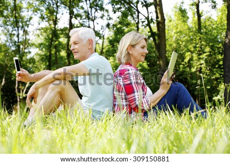 Portrait of senior couple sitting at the forest facing each other. Elderly woman holding hand digital tablet while senior man touching his mobile phone.