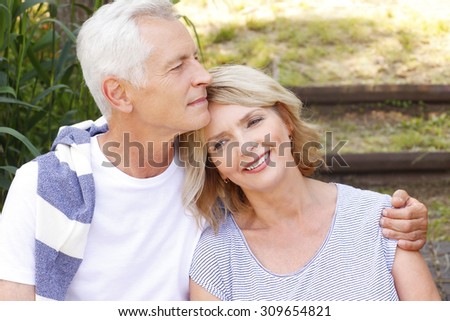 Close-up portrait of happy senior couple sitting at the lakeside. Elderly man hugs his wife while relaxing at nature.